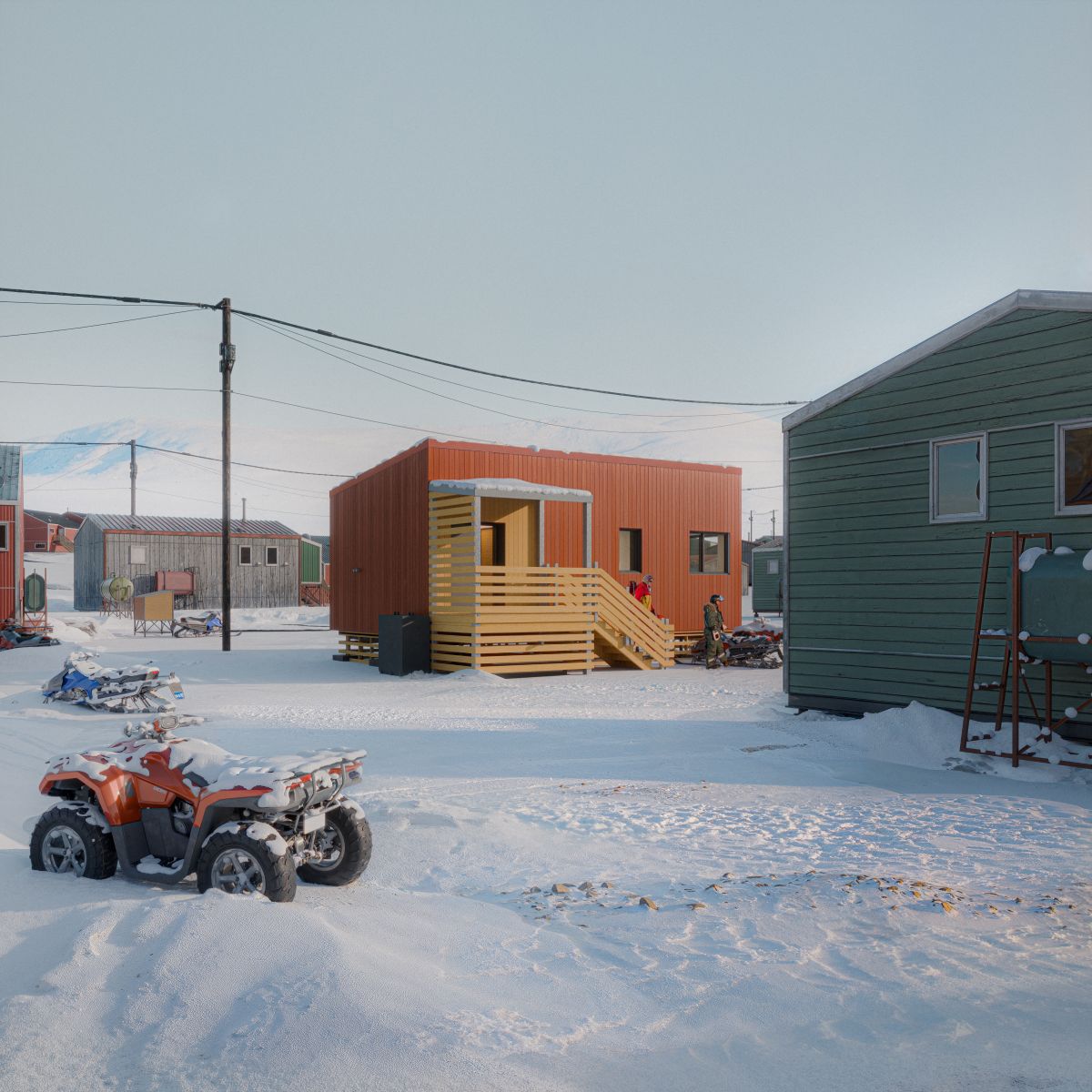 A 3D rendering of a  home with a corrugated metal facade and a covered entrance and set in a cold weather community. The structure features a raised wooden porch with horizontal slat railings and a staircase leading to the entrance. The home is surrounded by small buildings.